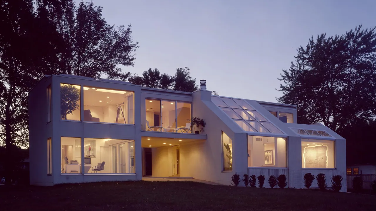 White modernist house at dusk with large glass walls and a sloped glass roof, rooms lit warmly inside, dark trees around it