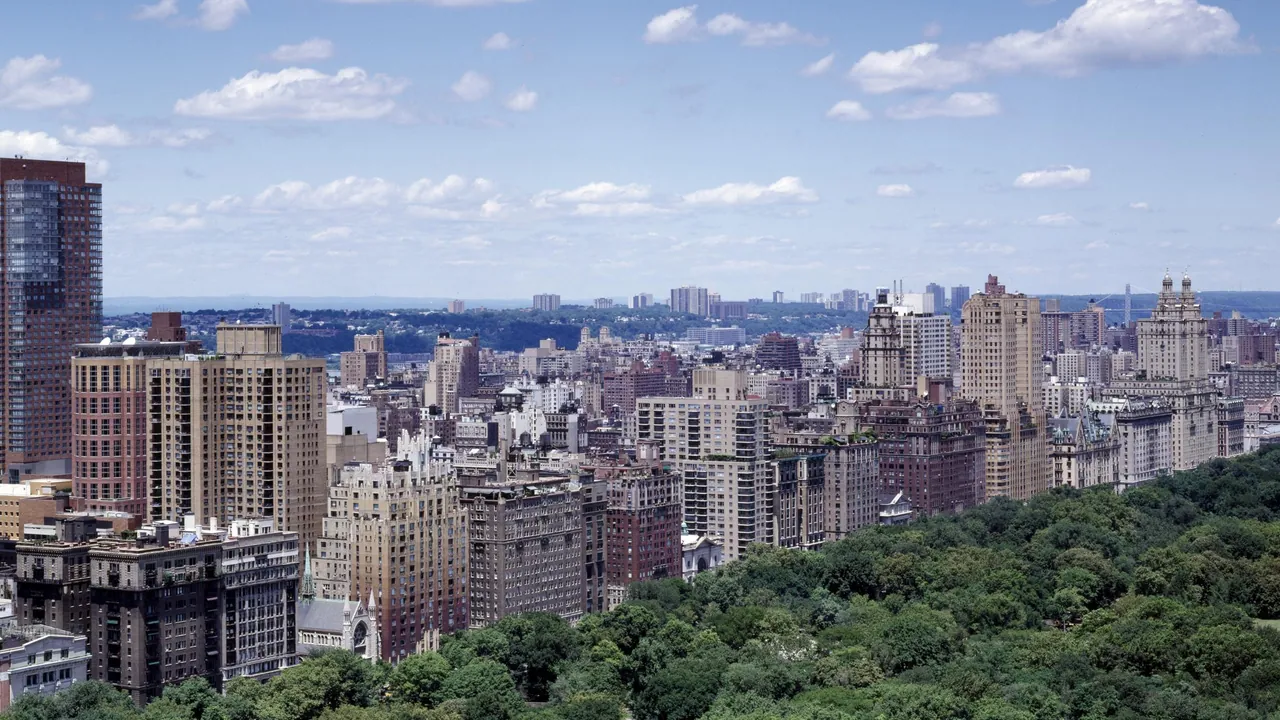 Aerial view of Upper West Side apartment towers lining the green treetops of Central Park under a blue sky with light clouds