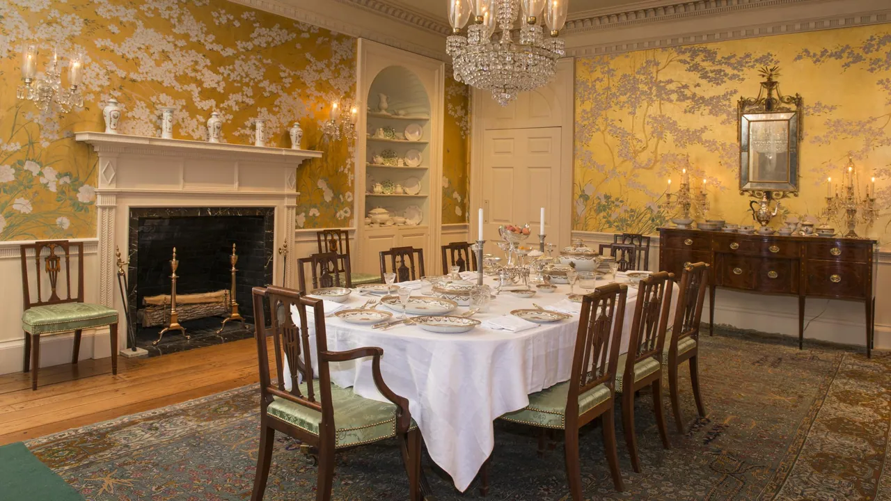 A formal dining room with hand-painted gold wallpaper, a crystal chandelier, a table laid with china and a mahogany sideboard with silver.