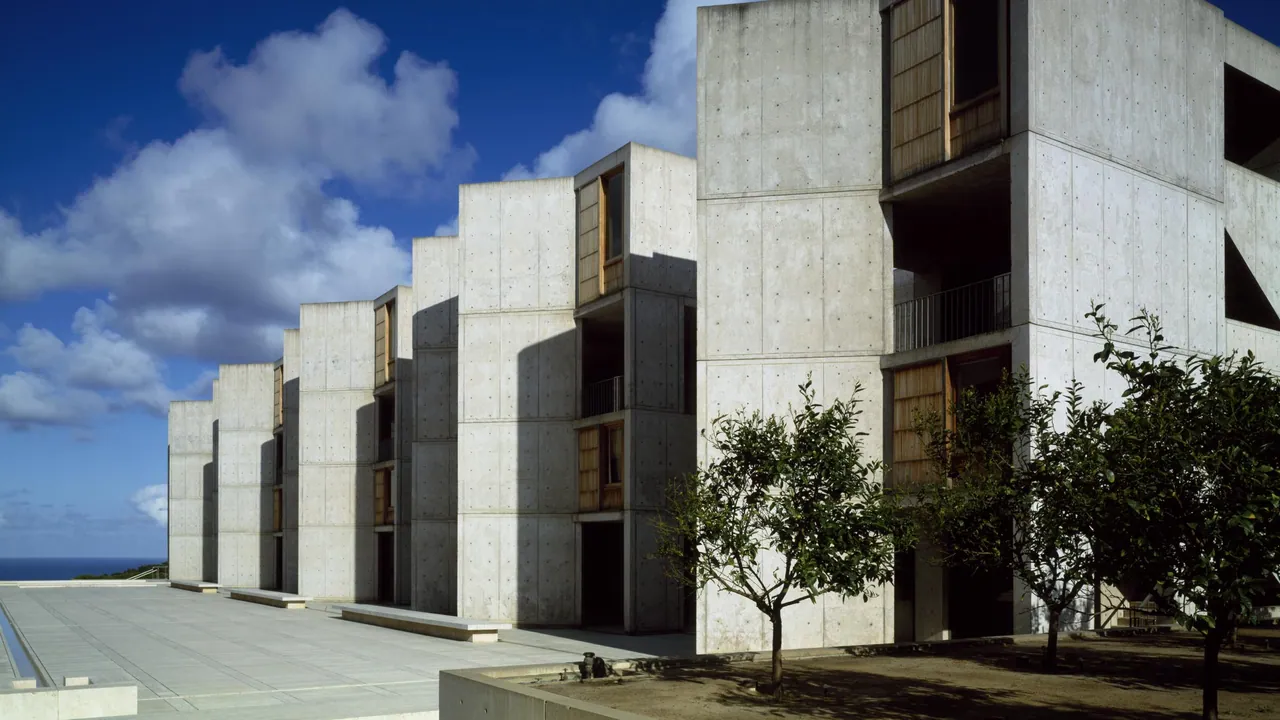 Concrete study towers with teak window panels step toward the ocean beside a stone plaza and young citrus trees under a blue sky.