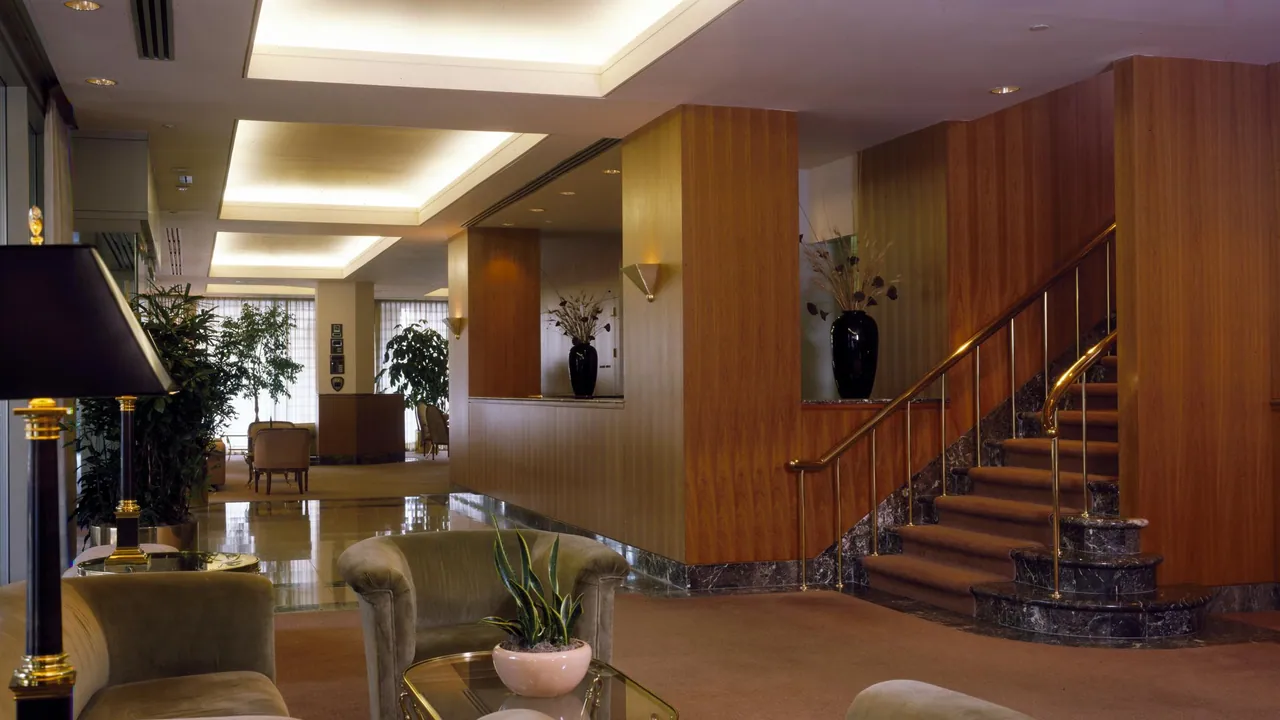 A quiet office reception lounge with upholstered armchairs, a glass table and brass lamp, warm wood-panelled walls and a marble-edged staircase with a brass rail