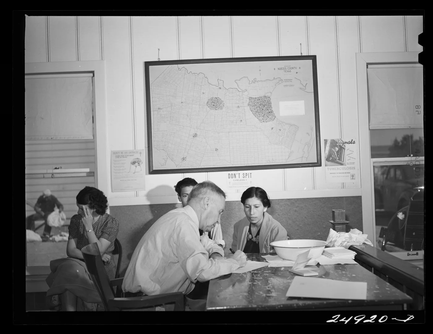 A doctor records a seated woman's medical history at a clinic table in 1942