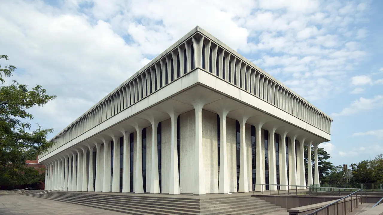 A white modernist building ringed by slender tapering columns under a deep fluted cornice, with broad steps and trees under a cloudy sky.