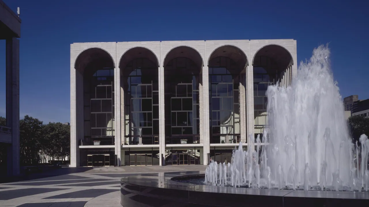 The arched glass facade of the Metropolitan Opera House at Lincoln Center behind a tall plaza fountain, set against a deep blue sky