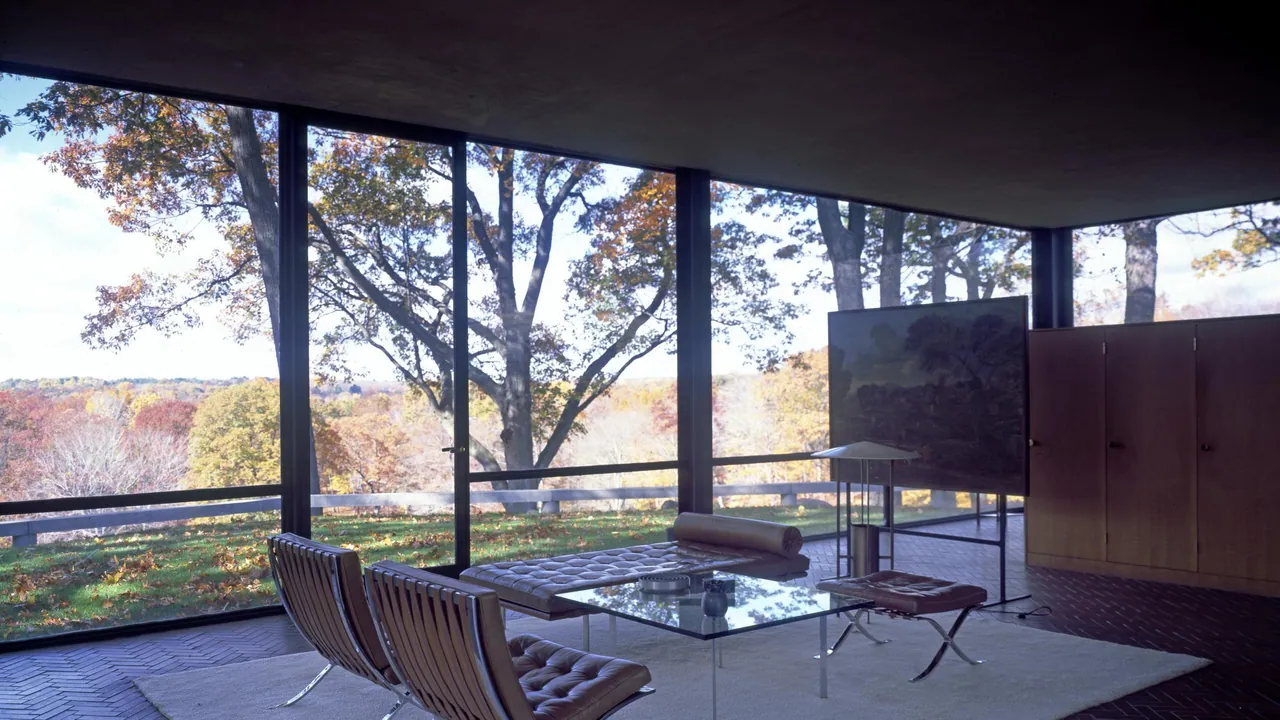 Leather Barcelona chairs and a glass table on a white rug in a glass-walled room looking out on autumn trees