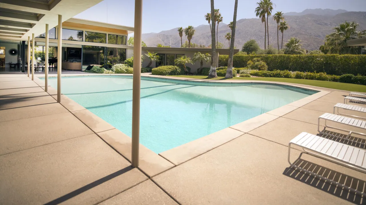 Mid-century house with long glass walls and a slim-post overhang beside a curving turquoise pool, white loungers, palms and desert mountains
