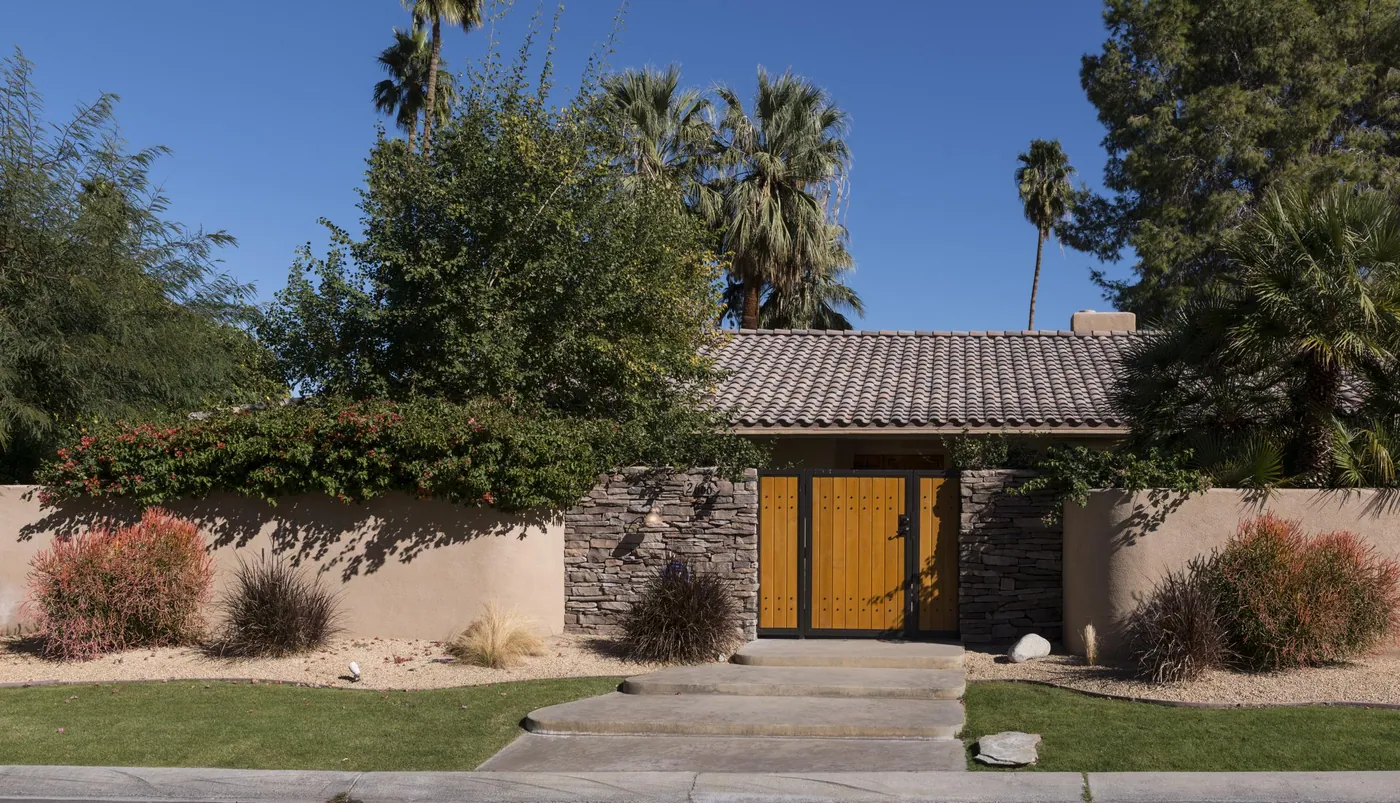 Single-story house with a tiled roof behind a low stucco wall, a stone entry and wooden gate, steps up from the lawn, palms and a blue sky