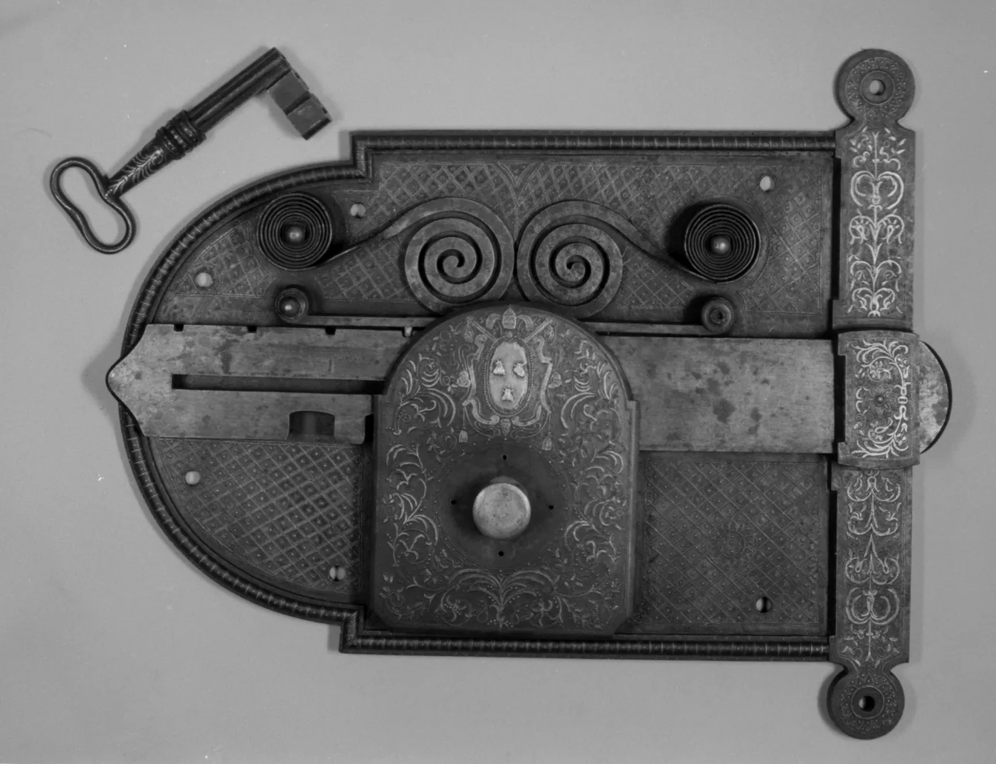 Black-and-white photograph of an engraved iron door lock with scrolled springs, a sliding bolt and a heraldic shield, with its key beside it