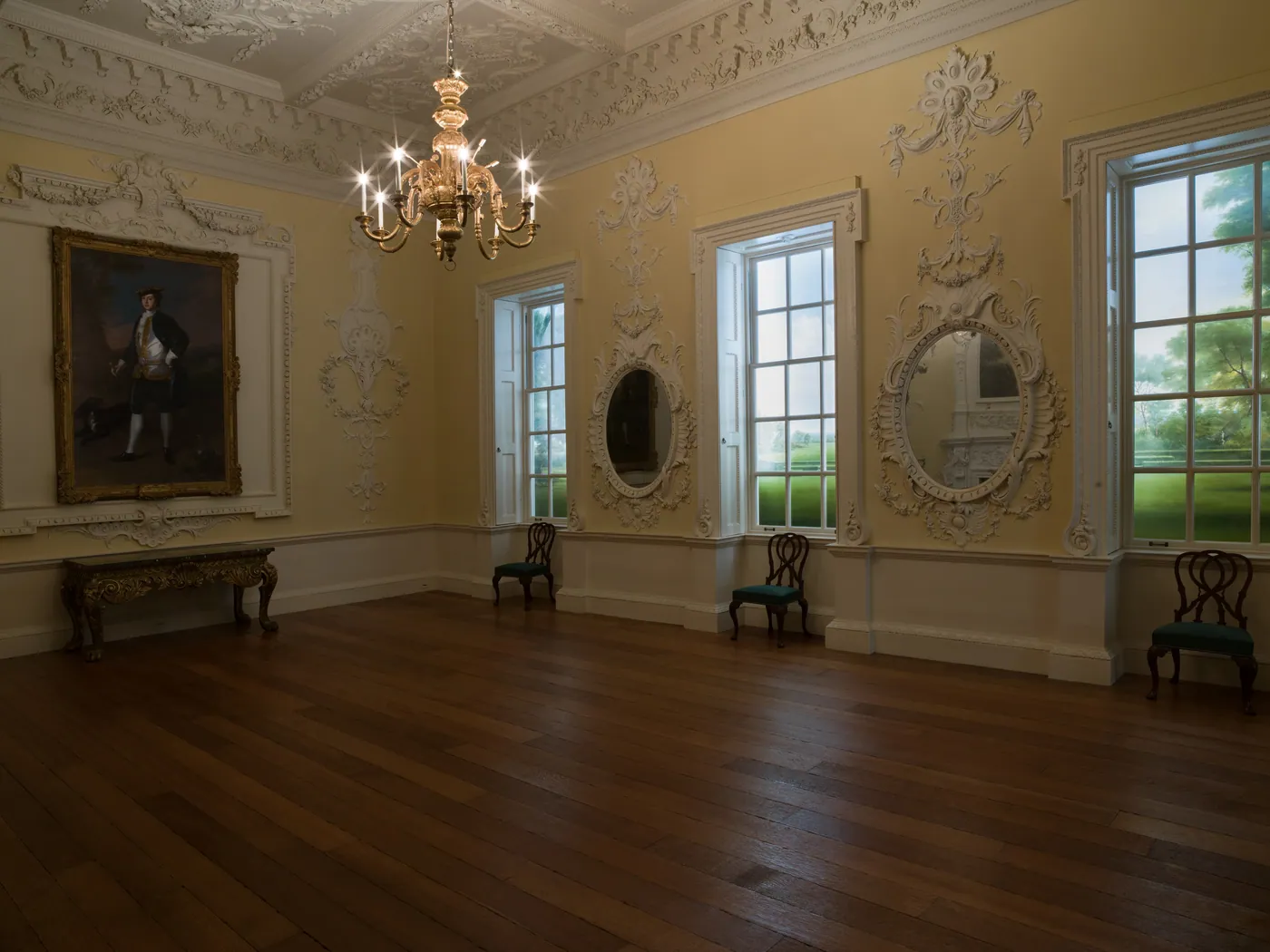 Empty Georgian room with plaster ornament, oval mirrors, tall sash windows, a lit chandelier, a portrait and a few chairs against the walls