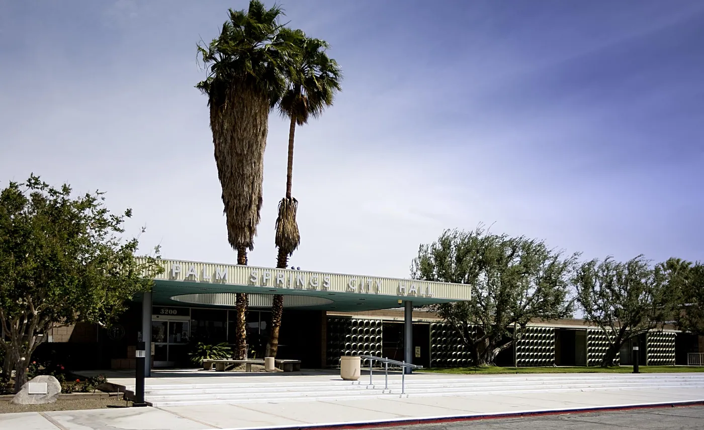 Low mid-century civic building with a flat curved entrance canopy, patterned block screens, tall palms and olive trees under a pale blue sky