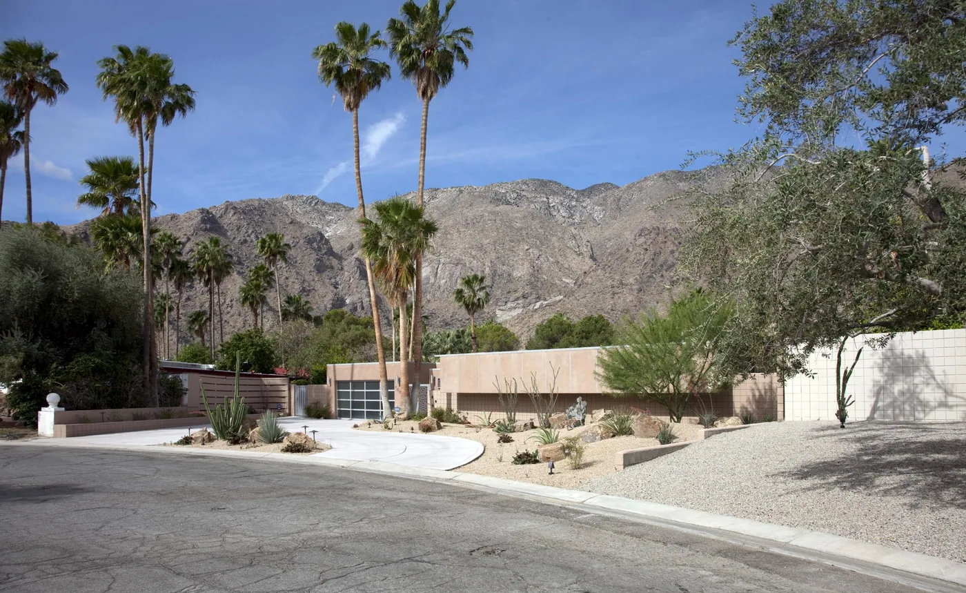 A low mid-century modern house with desert landscaping on a quiet curving street, tall palms and the San Jacinto Mountains behind