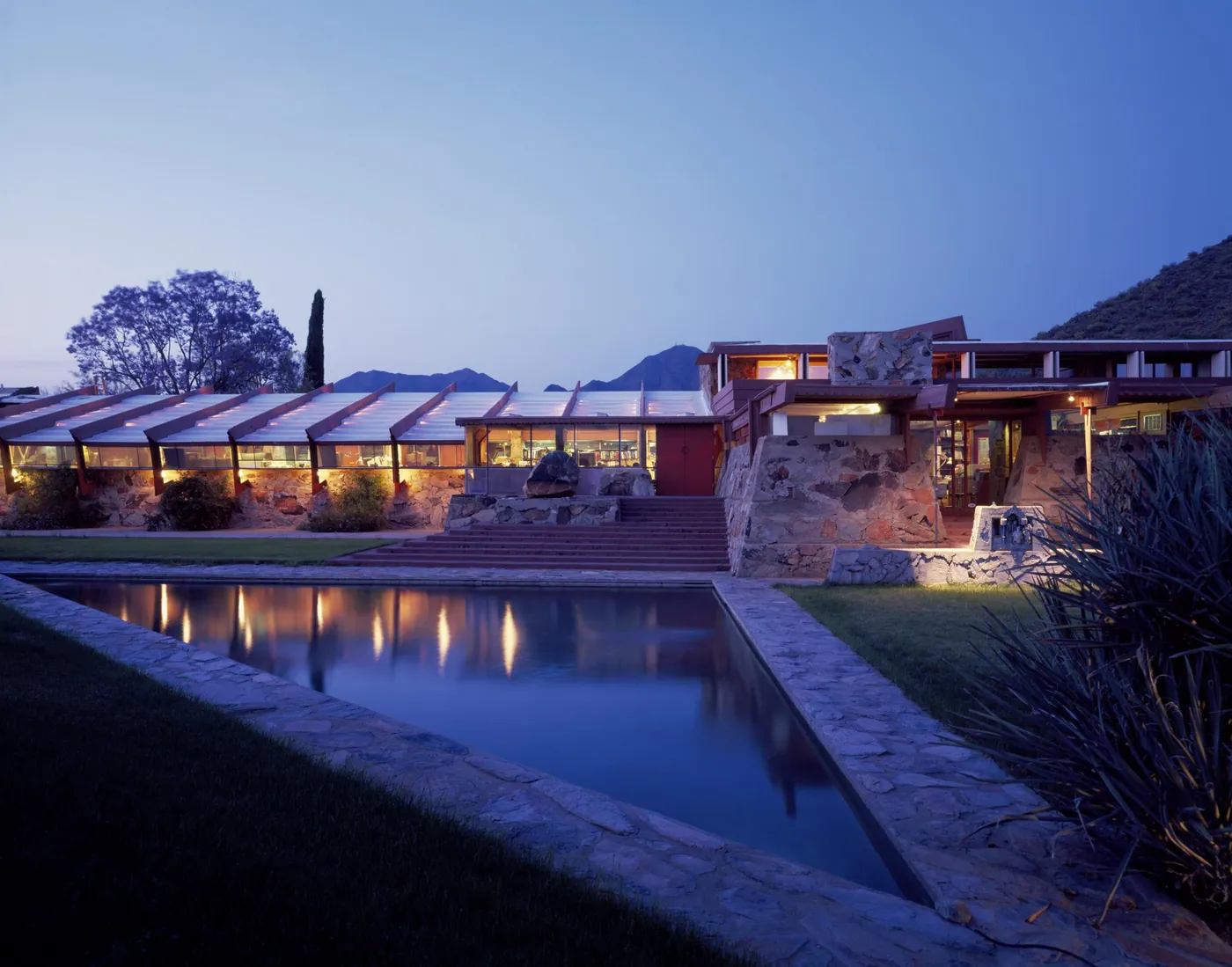 Frank Lloyd Wright's Taliesin West at dusk, its lit stone and timber rooms reflected in a long rectangular pool below desert hills