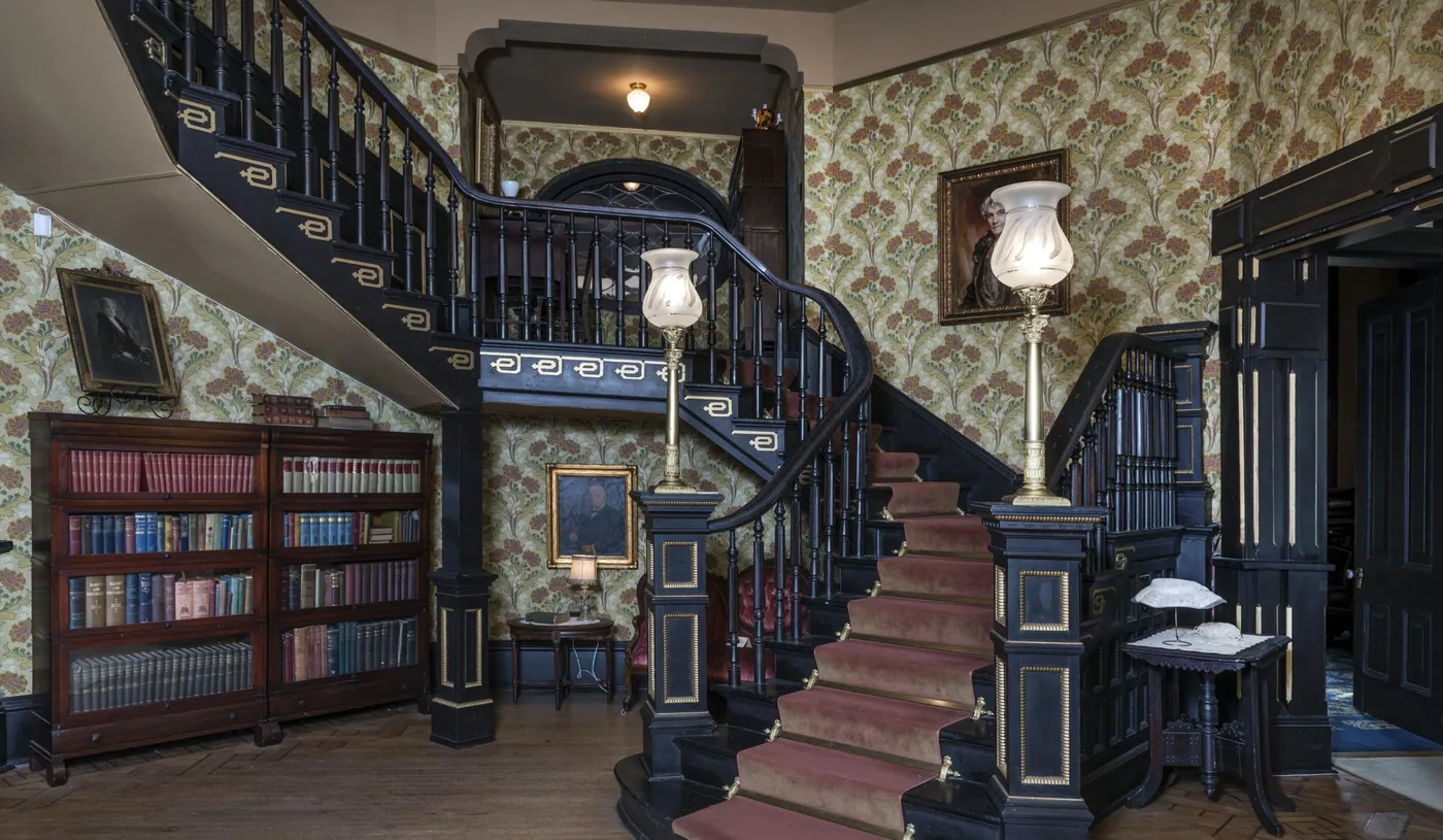 A carpeted Victorian staircase with dark banisters and tall glass lamps on its newel posts rises from a wallpapered hall lined with bookcases.