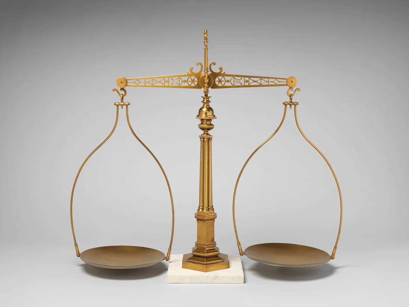 A gilt brass balance scale with a fluted column, pierced beam, and two shallow pans, standing on a white marble base