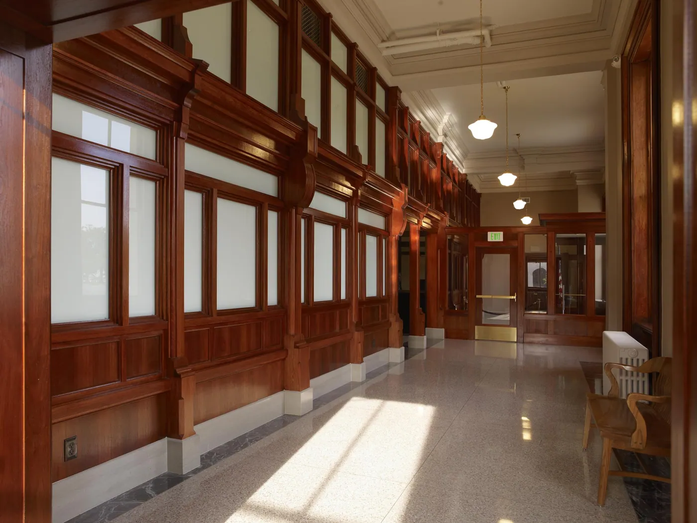 Sunlit courthouse corridor lined with polished mahogany panelling and frosted windows, pendant lamps overhead and a wooden bench by the wall