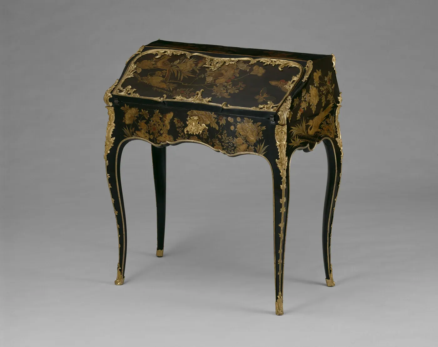 Black lacquer slant-front writing desk decorated with gilt flowers and birds, with gilt-bronze mounts and tall curving legs, on a grey ground