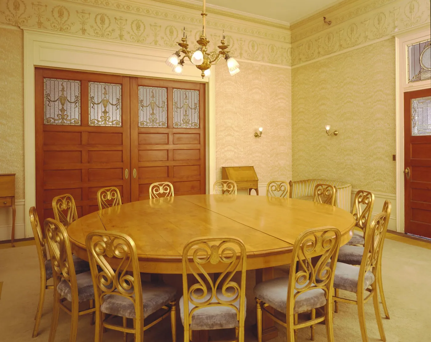 A round wooden table ringed by gilded chairs in a pale meeting room with carved doors, stained glass and a brass chandelier
