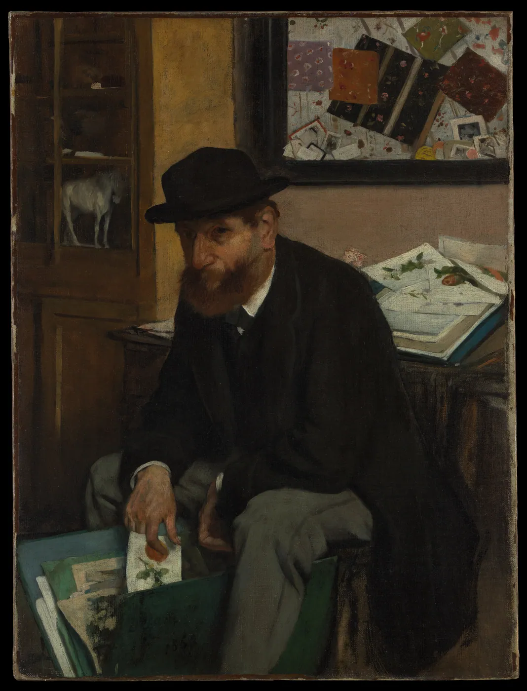A bearded man in a bowler hat sits leafing through a portfolio of prints, with more prints on a table and a pinboard behind him.