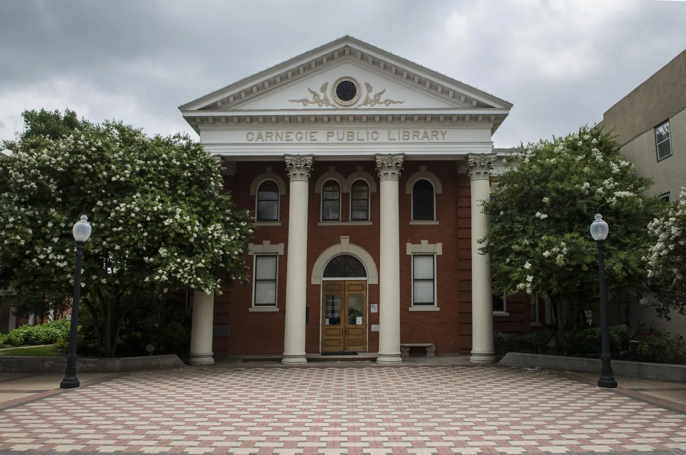Red-brick library with a white classical portico and Ionic columns, framed by flowering crape myrtle trees and lamp posts on a patterned plaza