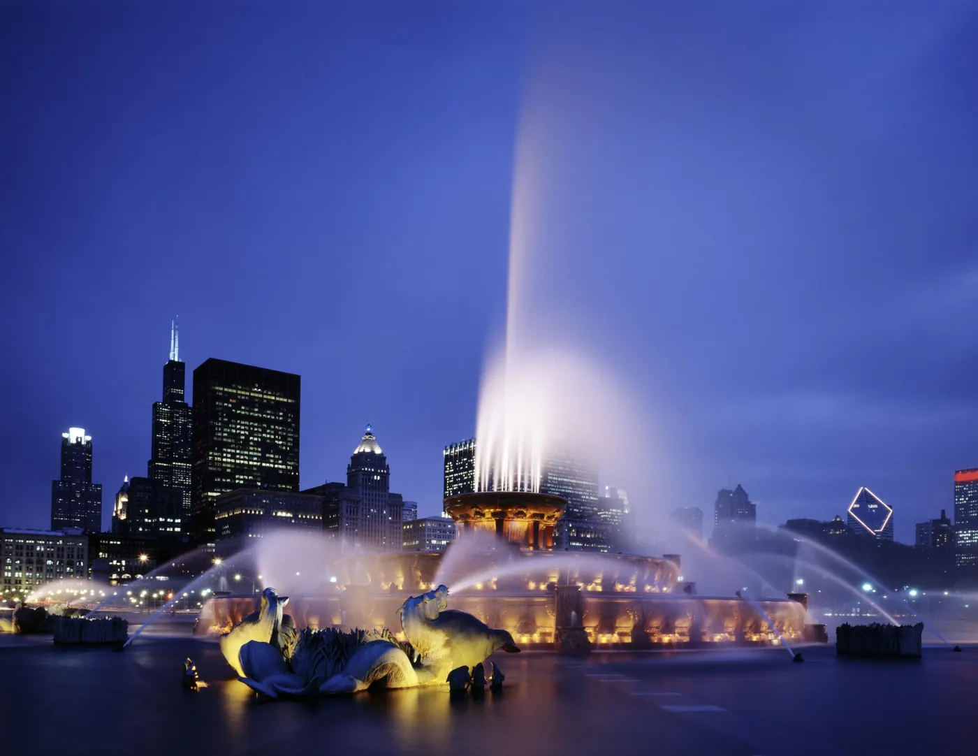 Illuminated fountain with bronze sea-horse sculptures sending tall jets of water into a blue dusk sky before the Chicago skyline