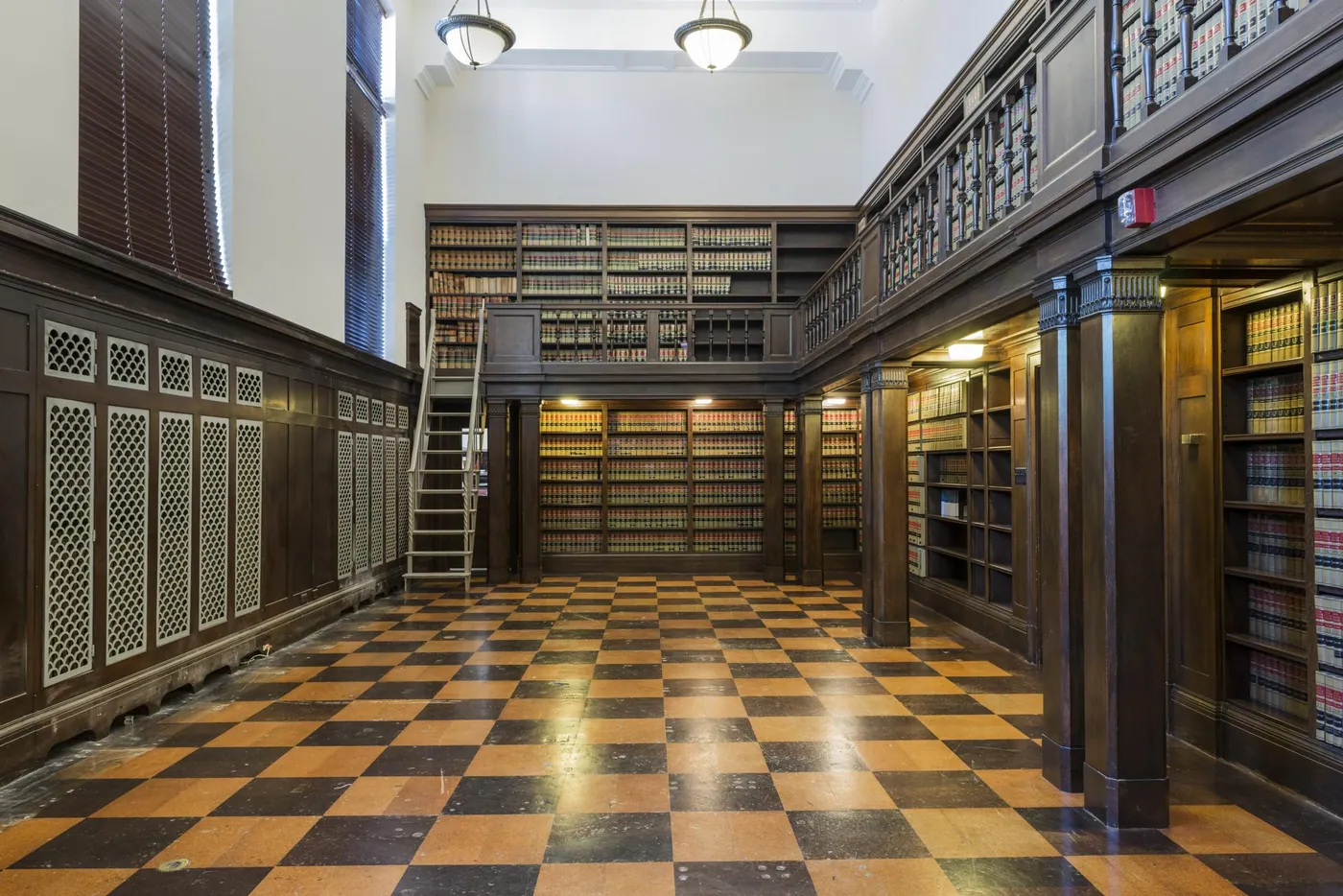 Two-storey law library with dark wood bookcases of bound volumes, a gallery railing, a ladder and a checkerboard floor