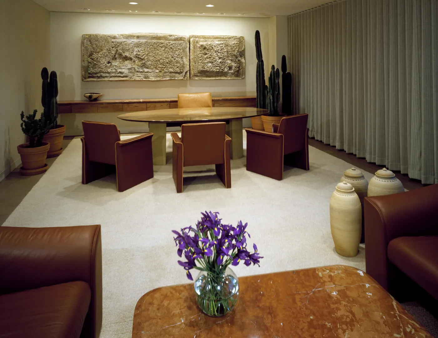 Quiet modernist conference room with leather chairs around an oval table, potted cacti, stone relief panels and a vase of purple irises