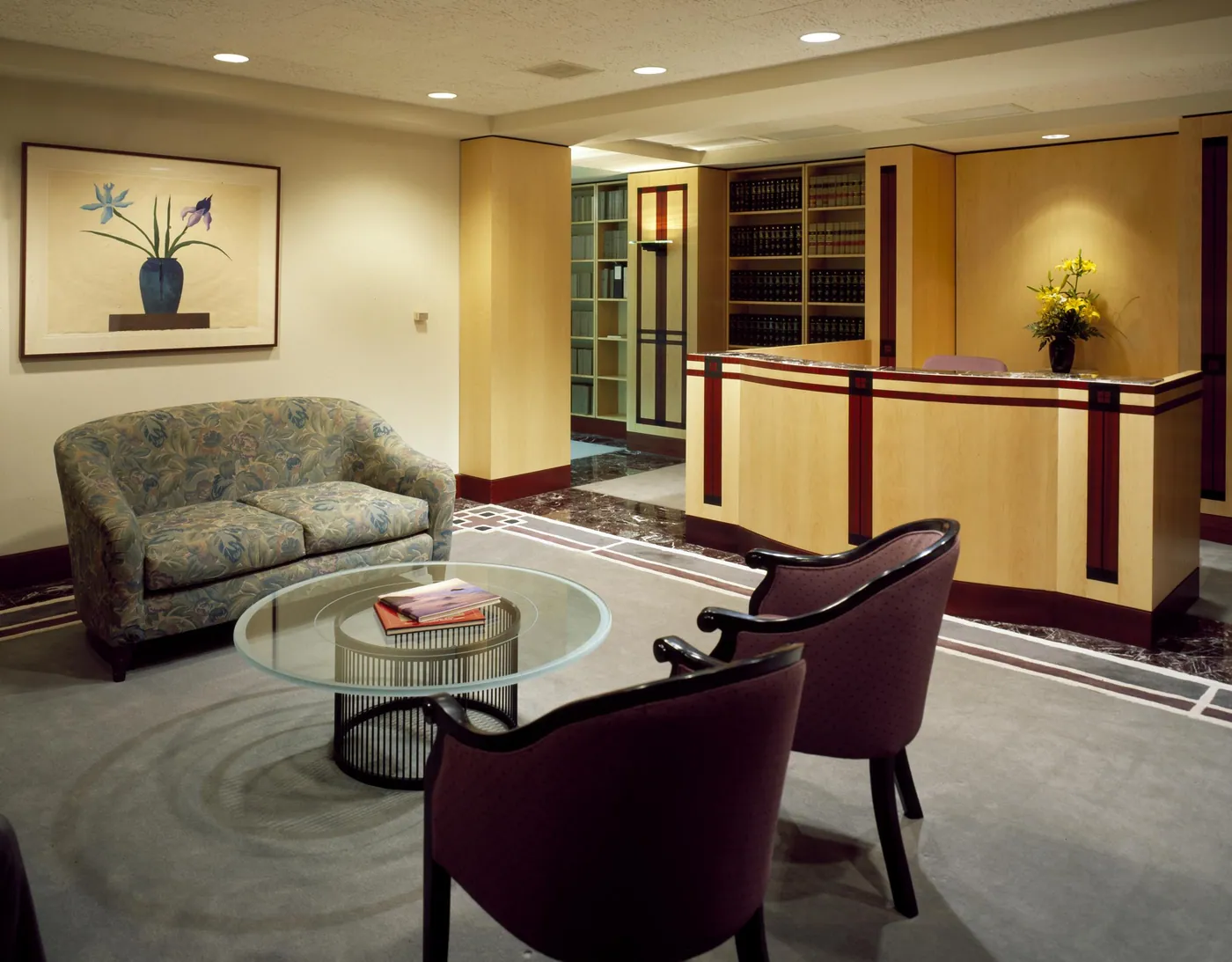 A quiet professional reception area with a sofa, glass coffee table, two armchairs, flowers on the front desk and shelves of law books beyond