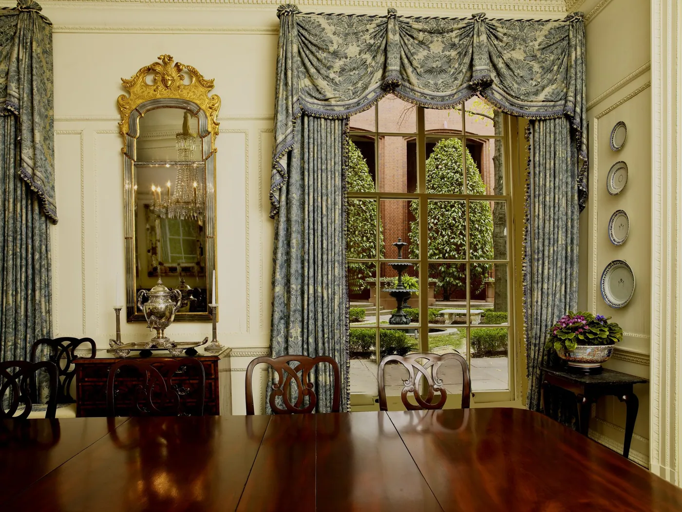 Formal dining room with a polished mahogany table and carved chairs, a gilt mirror, blue damask curtains and a window onto a courtyard fountain