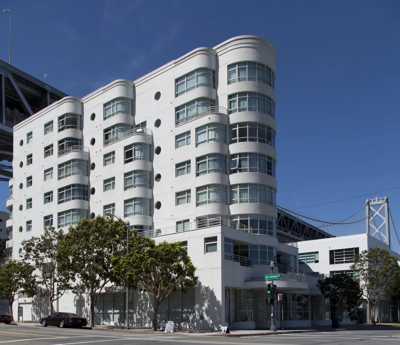 White Moderne-style apartment building with rounded bays, porthole windows and balconies against a deep blue sky, the Bay Bridge beside it