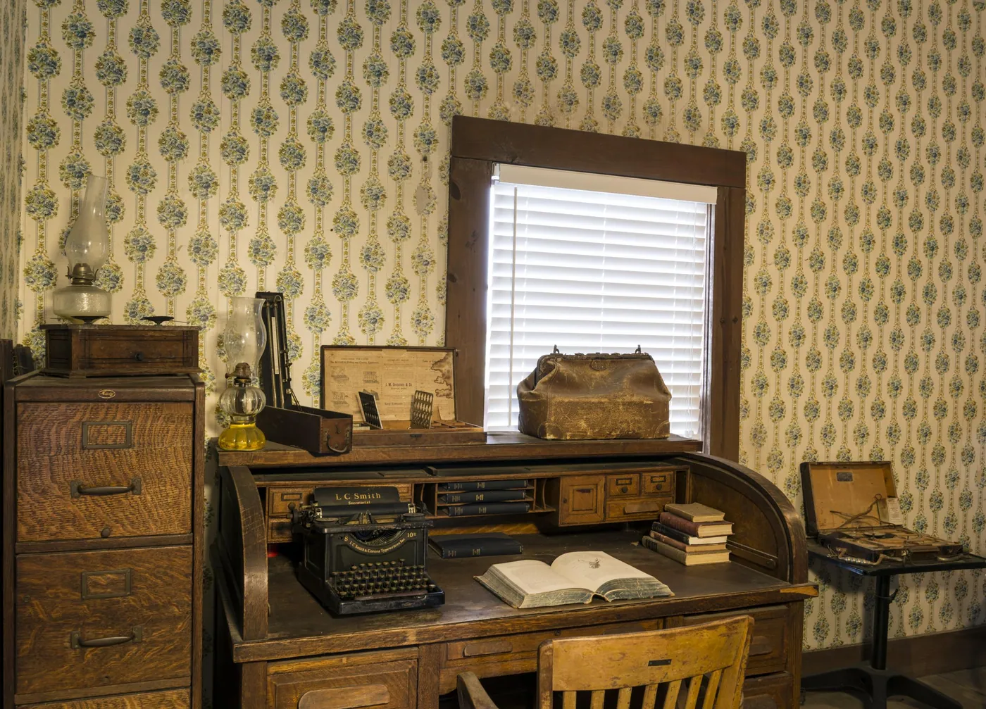 A wooden roll-top desk with an open book and an old typewriter, a leather doctor’s bag on top, beside a filing cabinet and patterned wallpaper.