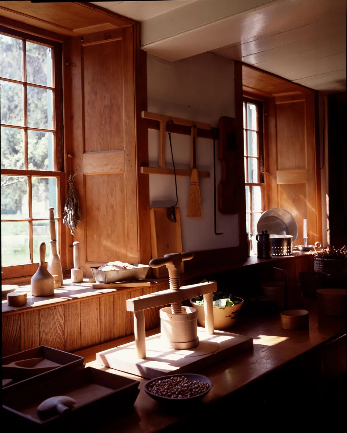 Sunlight falls through tall windows across a wooden Shaker kitchen counter holding a cheese press, a bowl of beans, greens and bread.