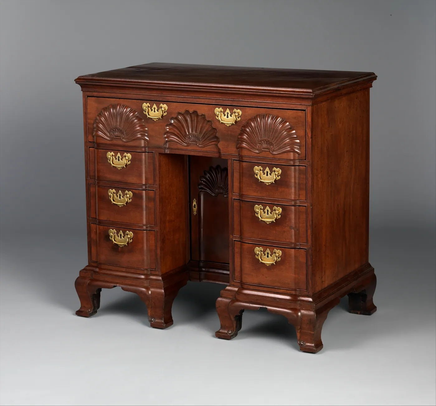 A mahogany kneehole bureau table with three carved shells across the top drawer, brass pulls and ogee bracket feet, on a grey backdrop