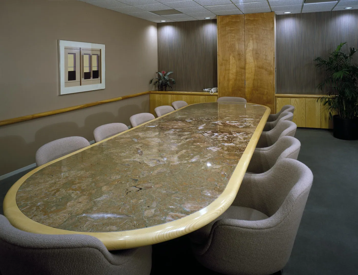 An empty conference room with a long oval marble table edged in pale wood, upholstered tub chairs, wood-paneled walls, and a potted palm.