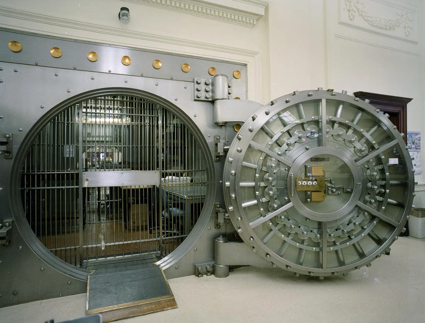 A massive round steel vault door swung open beside a barred inner gate, in a banking hall with cream moldings and brass bolts
