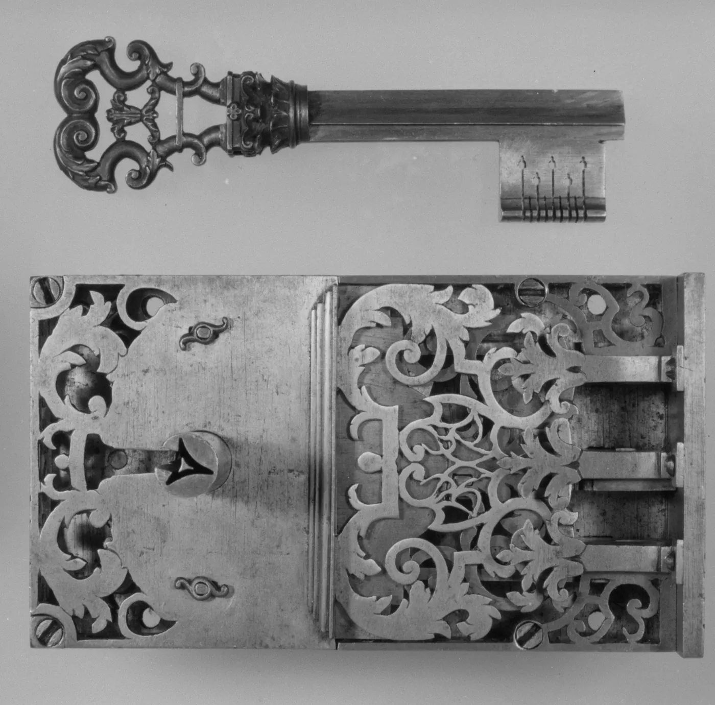 A black-and-white photograph of an ornate steel lock with pierced scrolling foliage and sliding bolts, shown below its large decorated key.