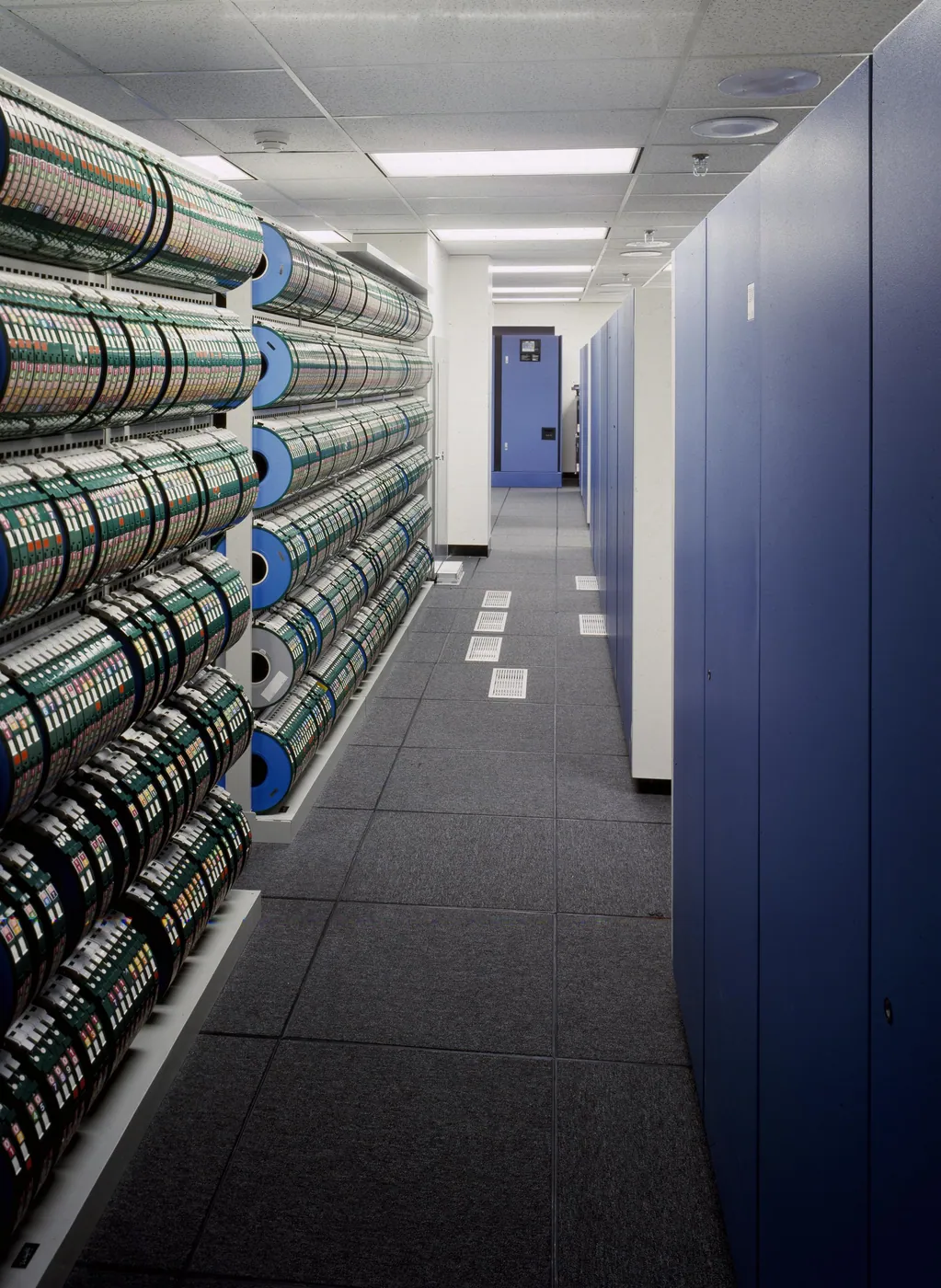 Long aisle of a data-storage room with racks of colour-coded tape reels on the left and tall blue cabinets on the right