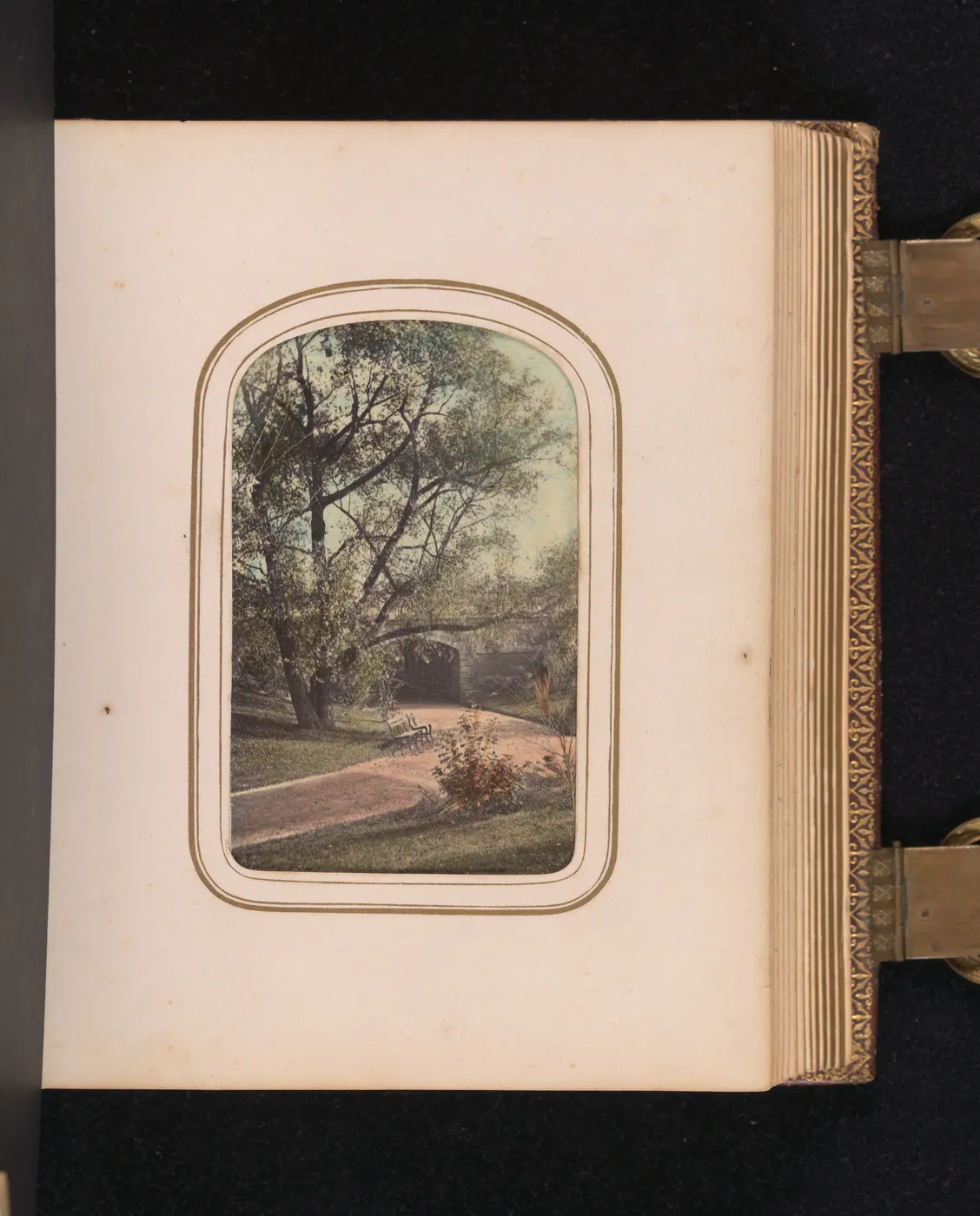 An open Victorian photograph album with gilt clasps, showing a hand-colored view of a Central Park path, trees and a stone arch