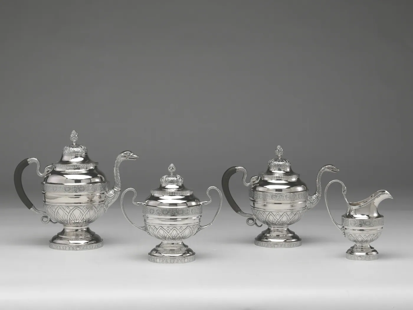 A four-piece silver tea and coffee service with swan-neck spouts and dark wooden handles, set out in a row on a soft grey background