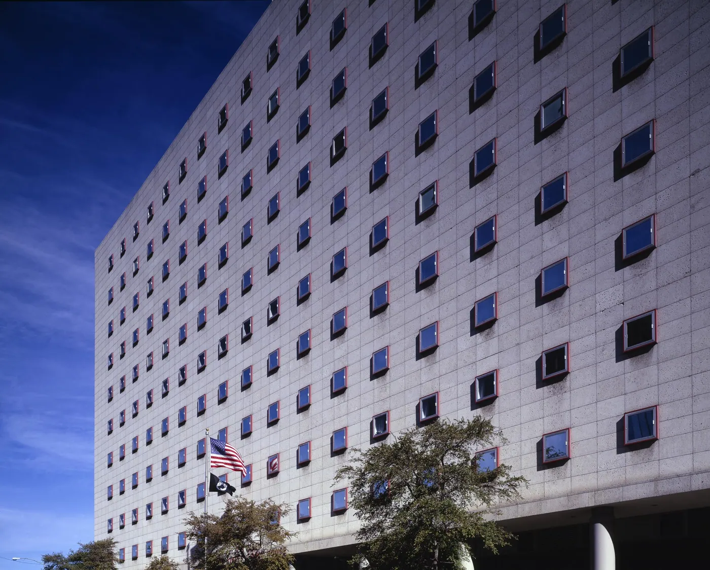 A 1962 modernist concrete courthouse with a grid of deep-set square windows under a deep blue sky, with flags and trees at its base