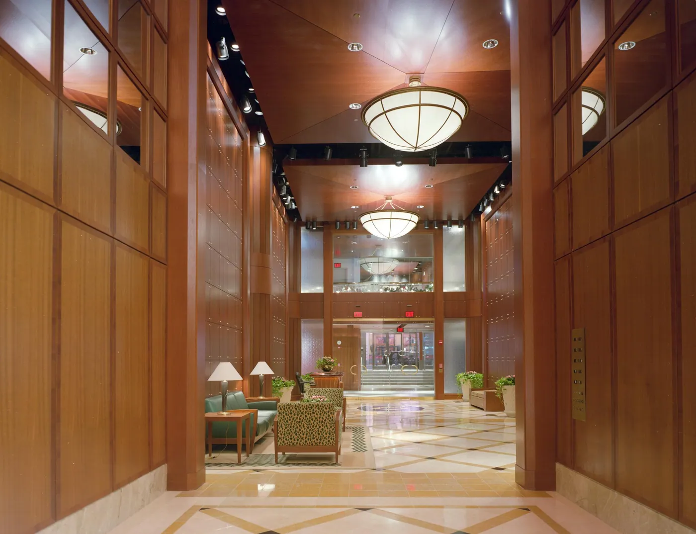 A tall office lobby lined with warm wood panels, globe pendant lights, a seating area with lamps and patterned armchairs, and a marble floor.