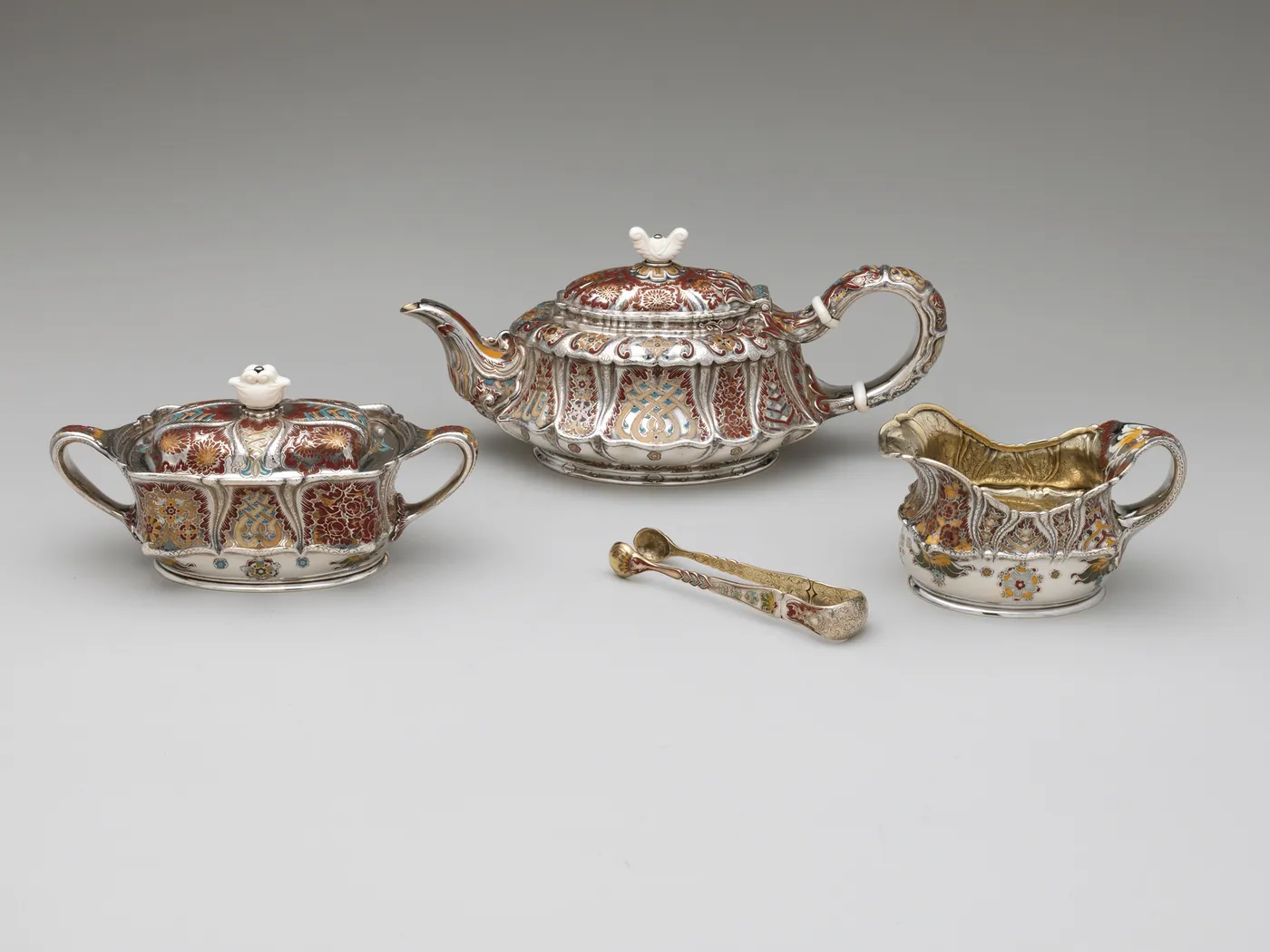 Silver tea service with teapot, covered sugar bowl, cream jug and tongs, decorated with red and gold enamelled patterns, on a grey ground
