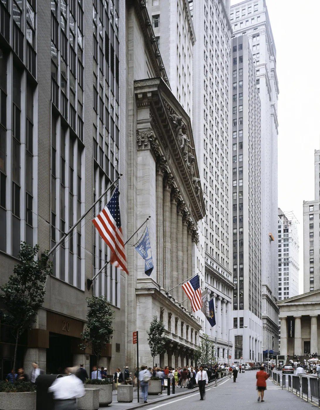 Looking down Wall Street past the columned New York Stock Exchange hung with flags, toward Federal Hall, with pedestrians in the street.