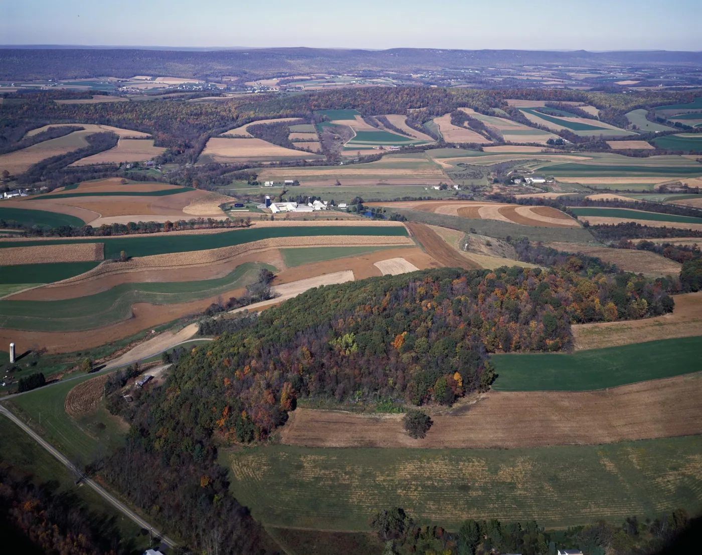 A patchwork of autumn fields, curving contour strips, farmsteads and a wooded hill seen from the air, stretching to a hazy ridge