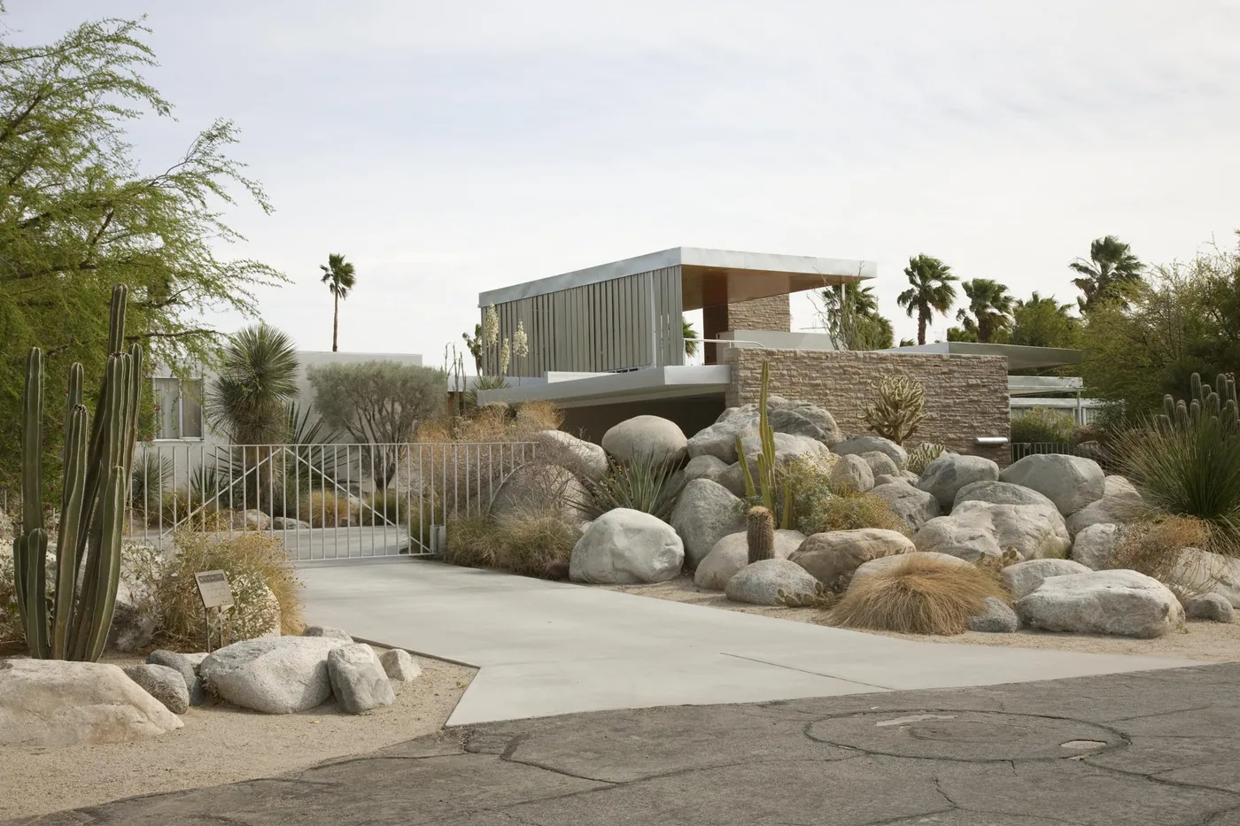 A flat-roofed modernist house with a louvered upper pavilion and stone walls, fronted by large desert boulders, cacti and a gated drive