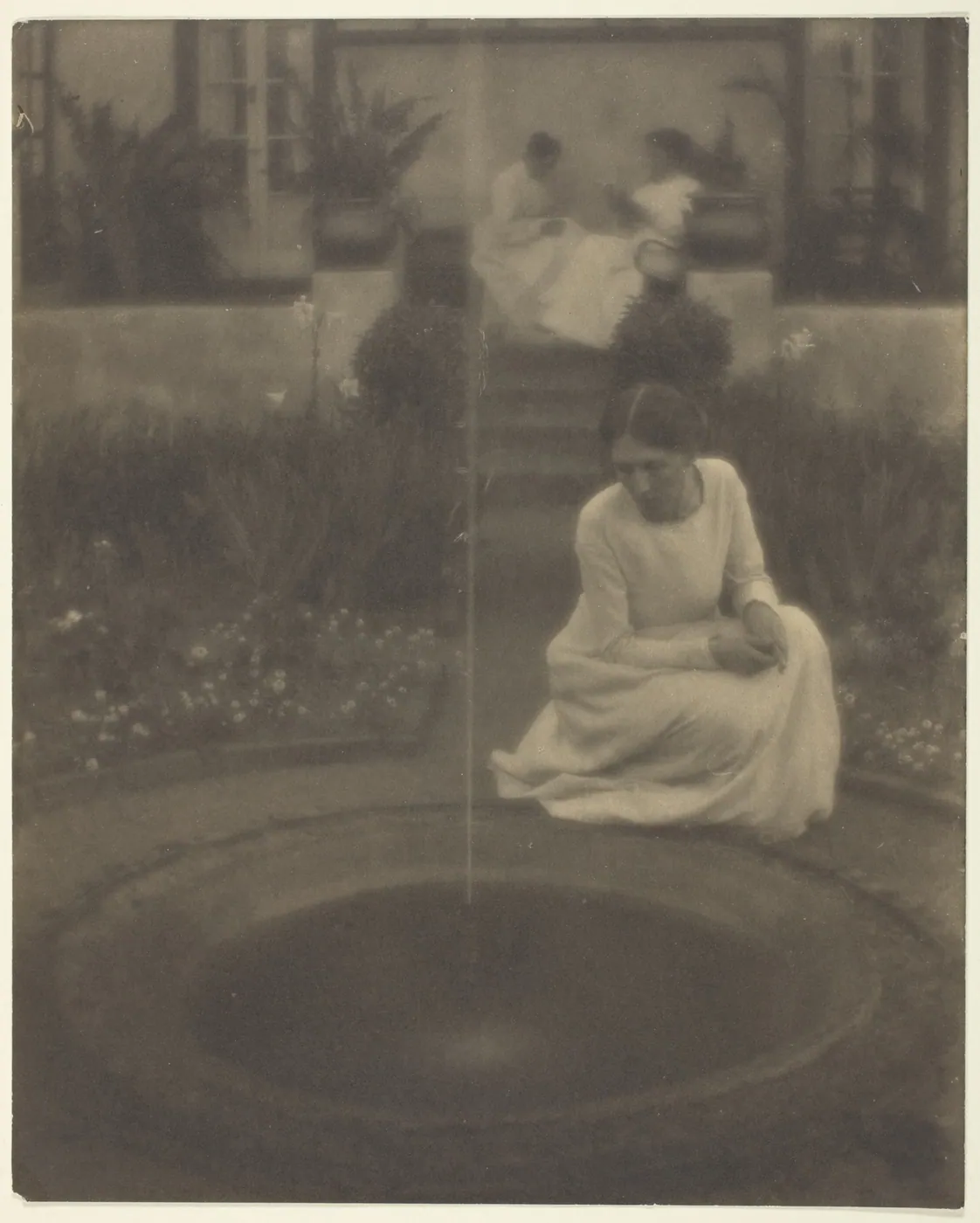 A woman in a white dress crouches beside a round garden pool, studying a single thin jet of water, while two figures sit on the terrace steps behind.