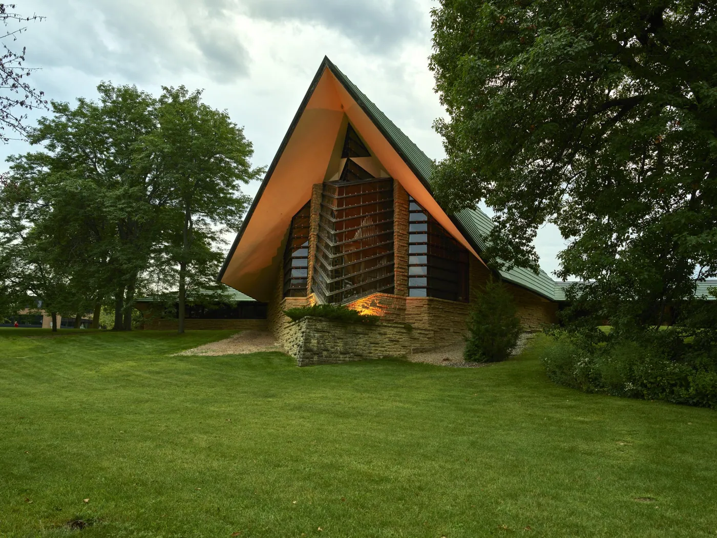 The copper-roofed prow of a stone and glass meeting house rises among tall trees above a broad green lawn, its windows softly lit