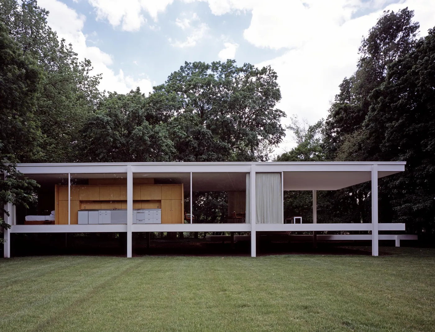 Glass-walled mid-century house with a white steel frame raised on slender columns above a green lawn, set against tall trees