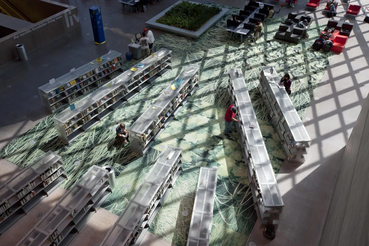 Overhead view of the Seattle Central Library floor, low book stacks on a green patterned carpet under a sunlit diamond grid of shadows
