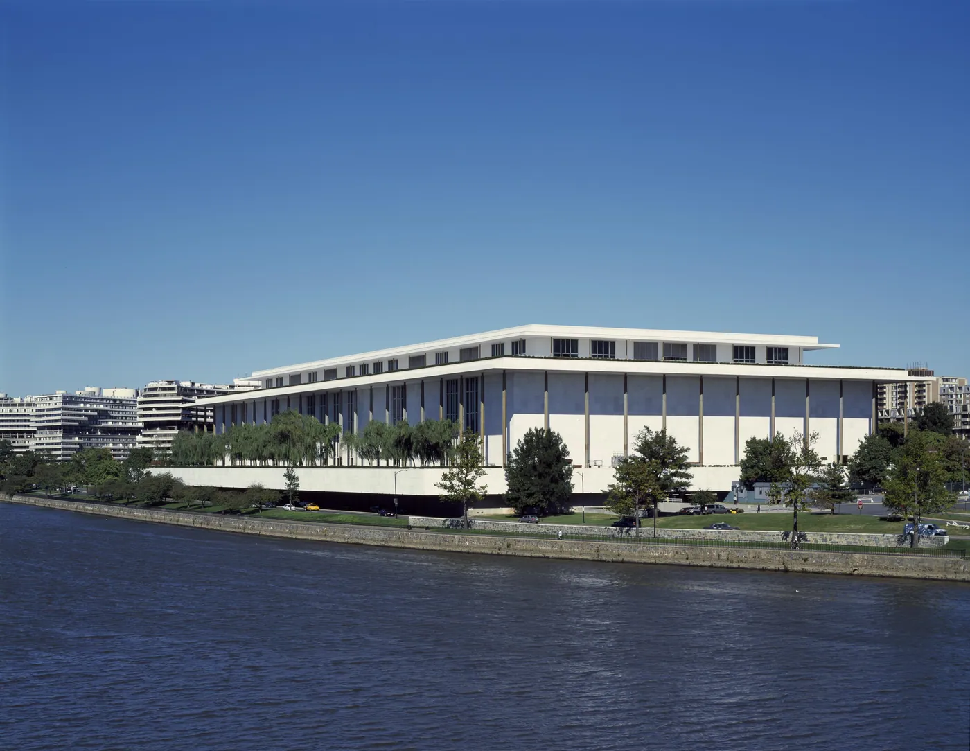 White marble Kennedy Center with a flat overhanging roof and slender columns, set along the Potomac River beneath a clear blue sky.