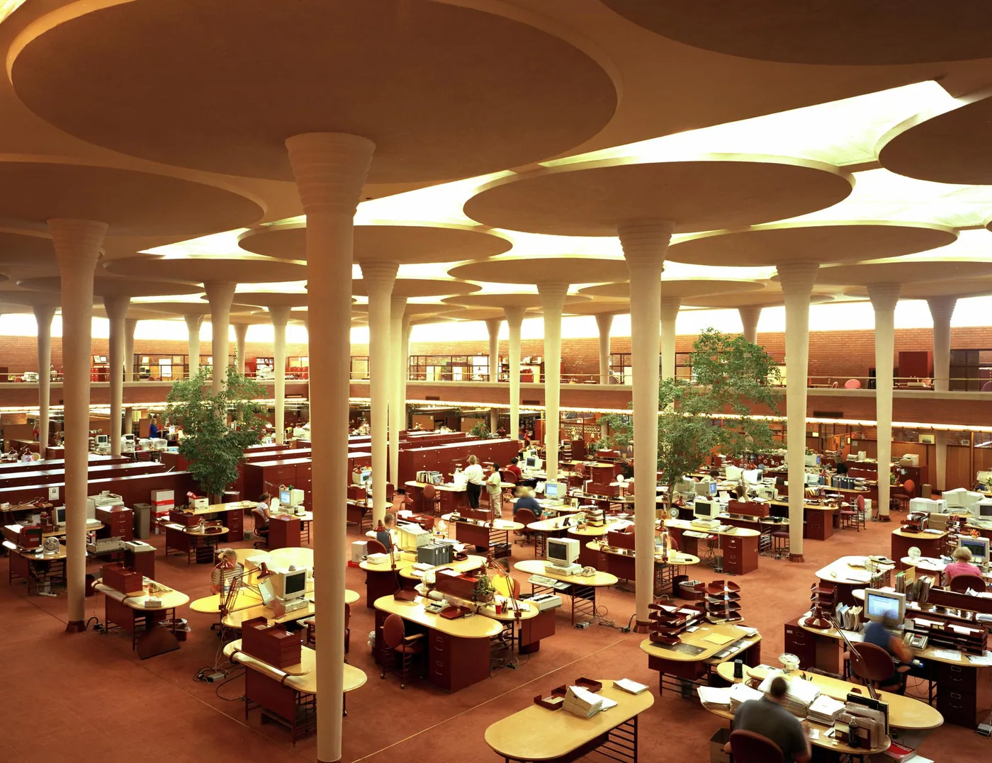 A vast skylit office hall with slender mushroom-shaped columns, rounded desks and warm brick-red floors, staff working at small distance